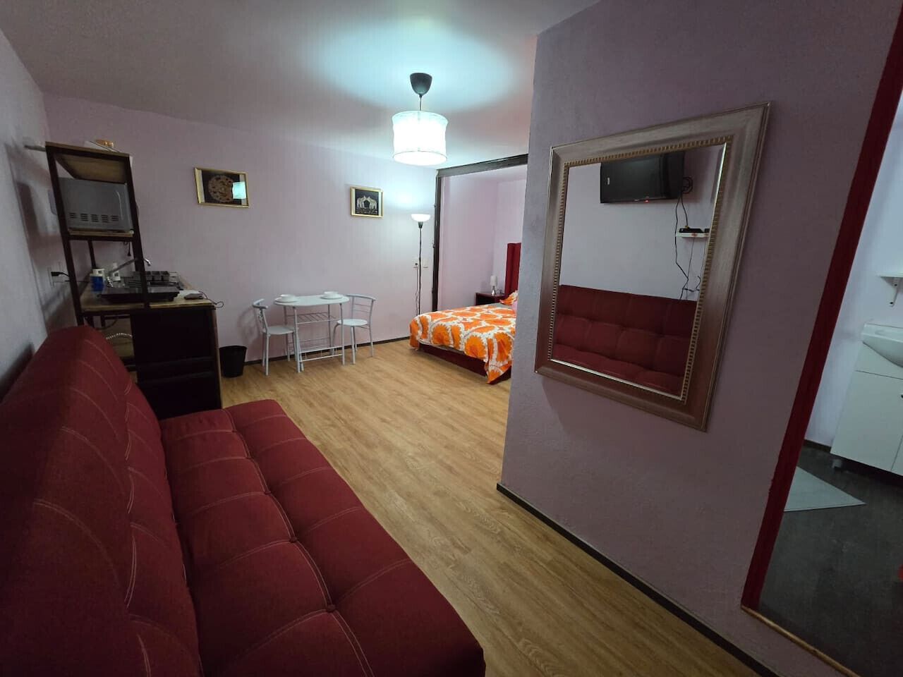 Una habitación con un sofá rojo, una cama con colcha naranja, un escritorio con computadora, una silla y un espejo en la pared.