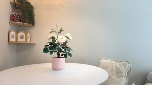 A round white table with a pink pot containing white flowers, a beige cushion, and a wall-mounted shelf with a plant and framed pictures. - Design Studio in Navigli | Wi-Fi & Self Check-in (Milano)