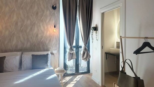 A bedroom with a bed, a window with curtains, a hanging light fixture, and a coat rack with a bag. - Design Studio in Navigli | Wi-Fi & Self Check-in (Milano)