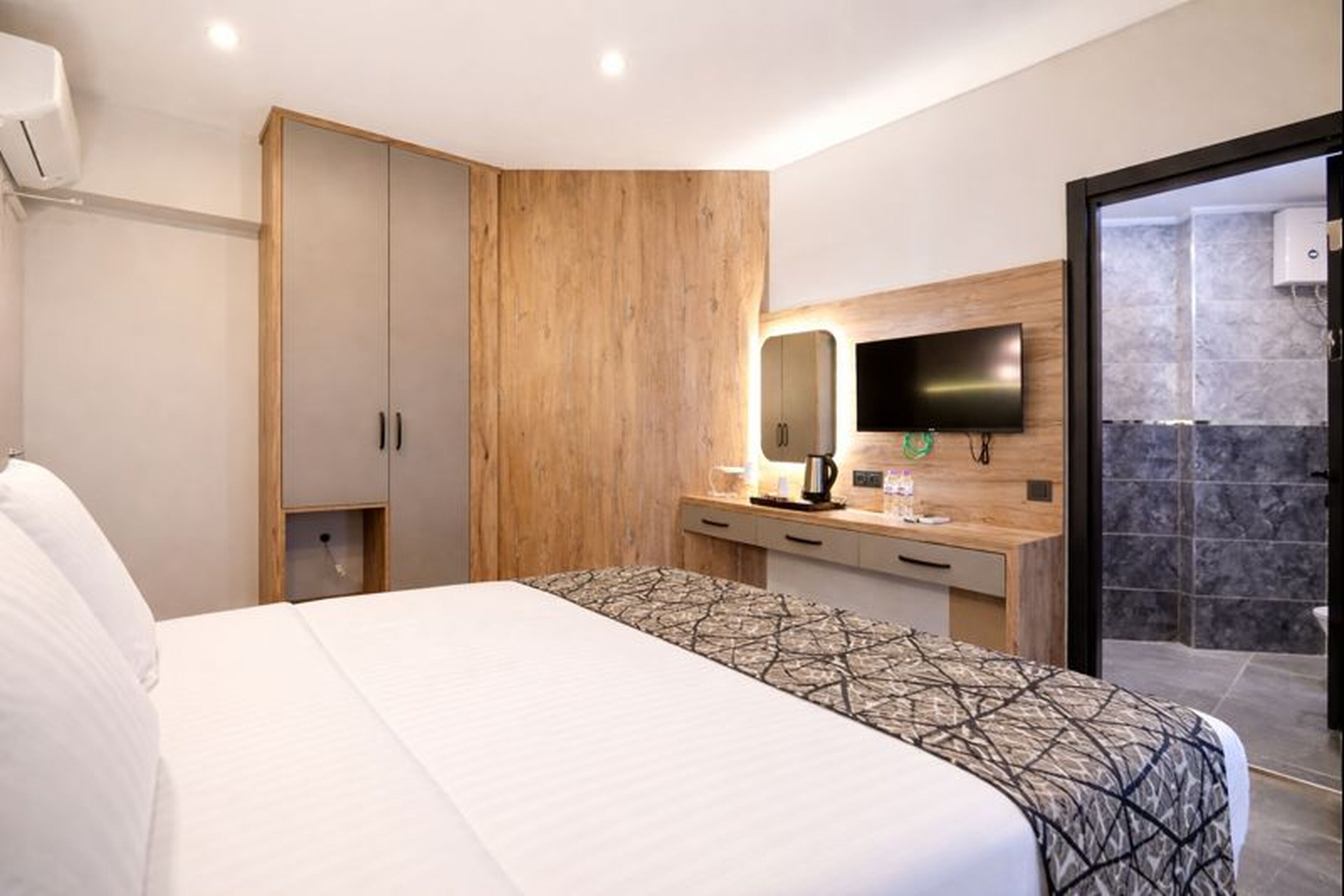 Une chambre d’hôtel moderne avec un grand lit, une armoire en bois, un petit bureau avec un miroir et un téléviseur fixé au mur.
