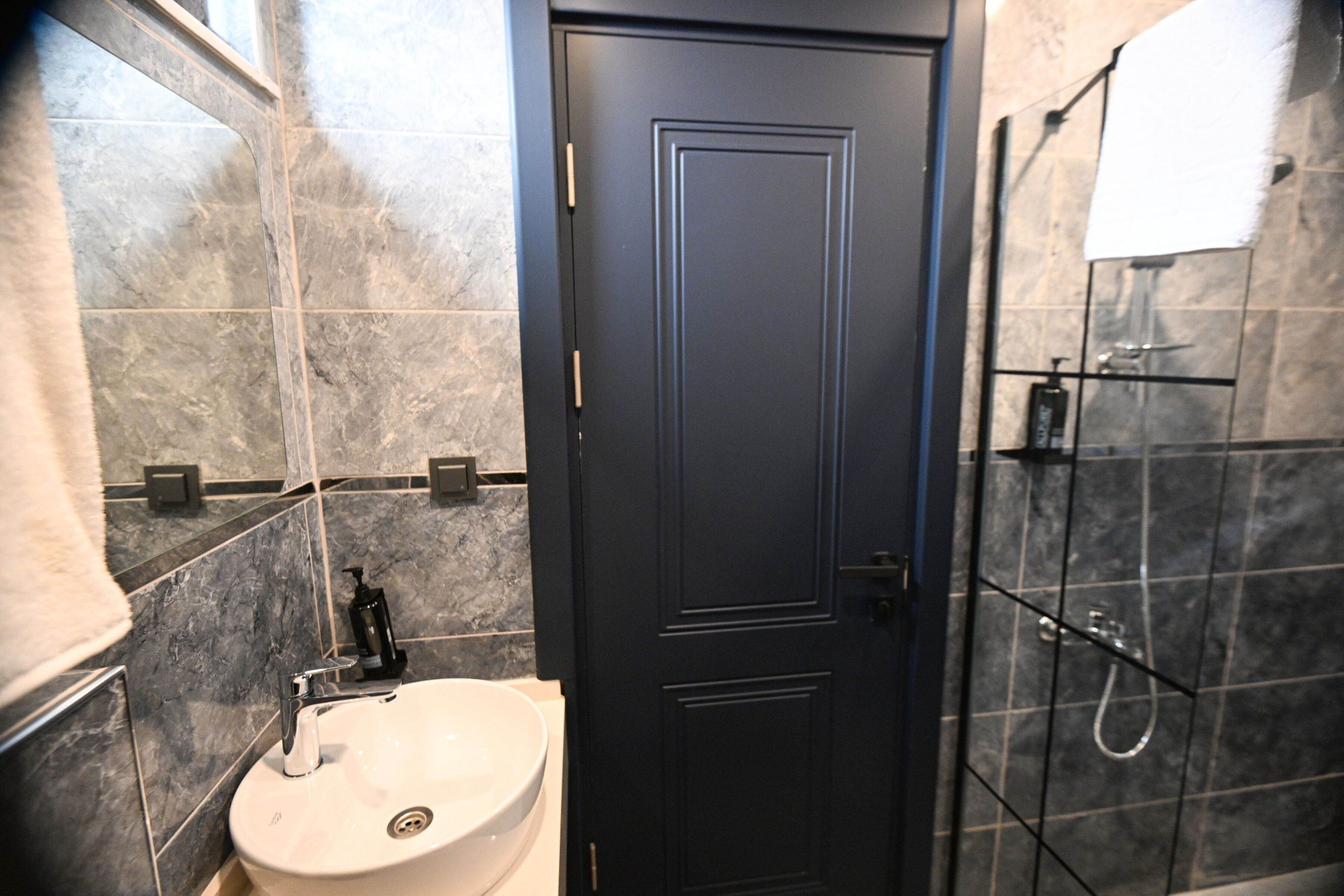 A bathroom with a white sink, dark tiled walls, a black door, and a shower area.