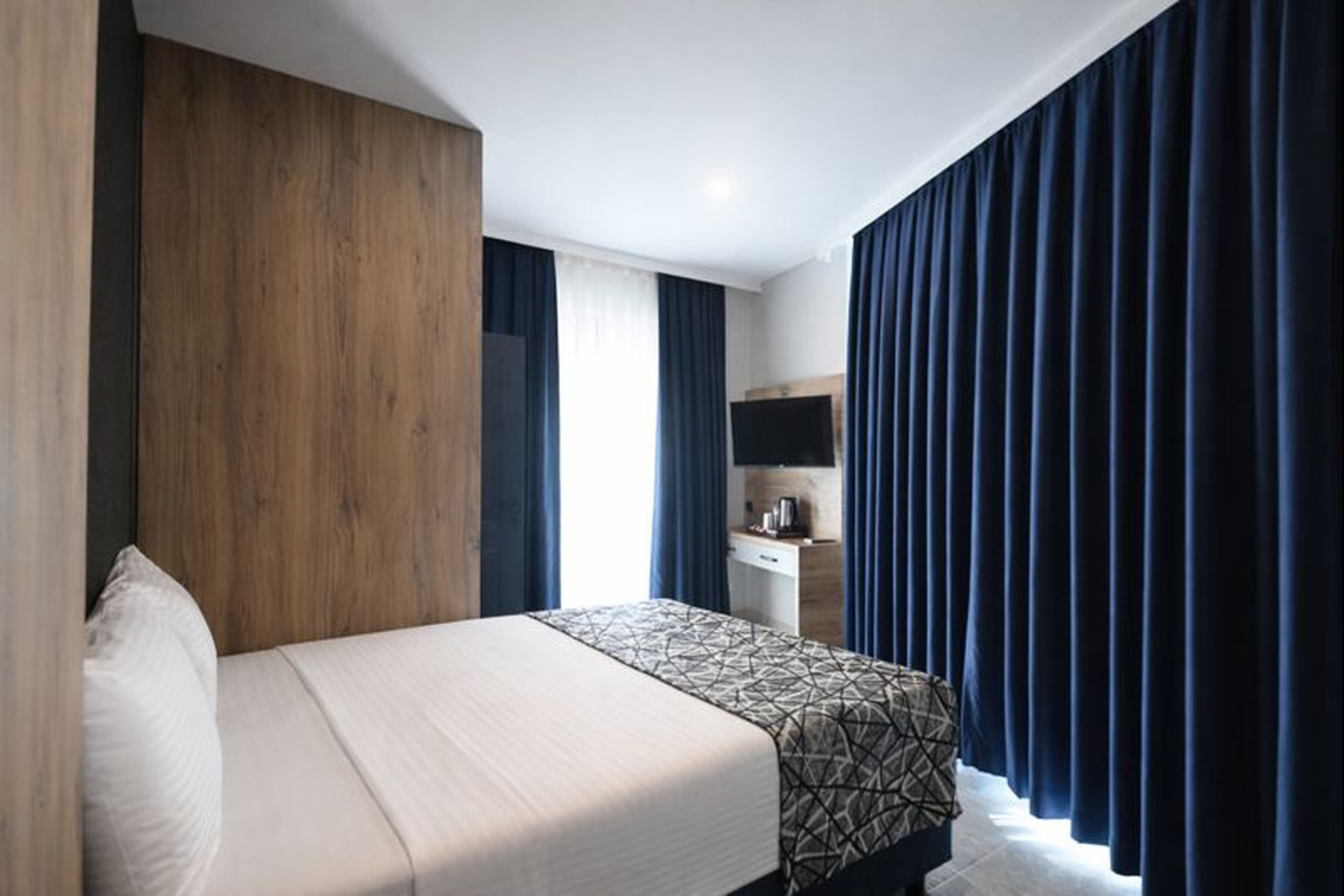 Una habitación de hotel con cama, cabecero de madera, escritorio y televisor de pantalla plana, además de cortinas oscuras.