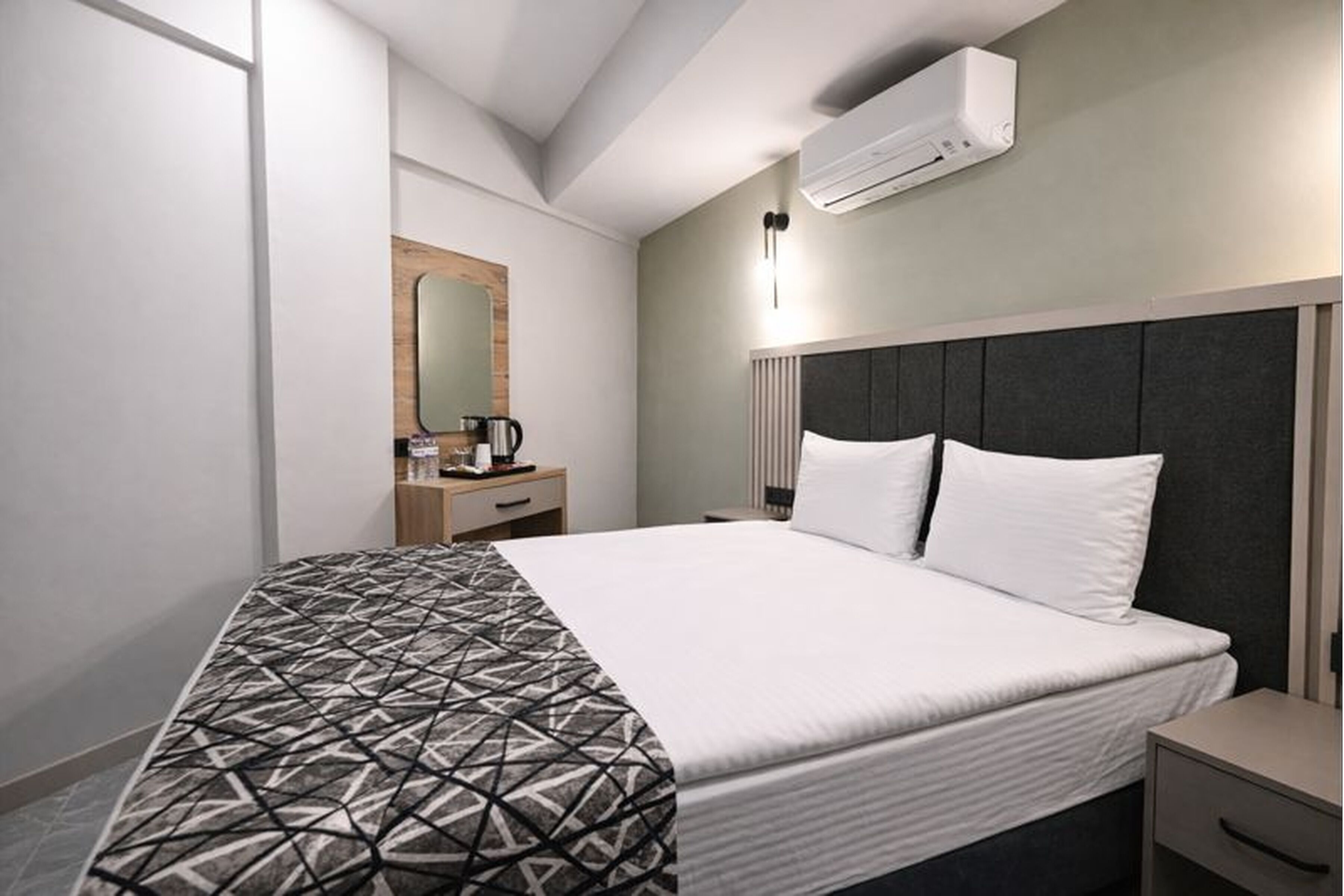 A modern hotel room with a large bed, a bedside table, a nightstand, a mirror, and an air conditioning unit.