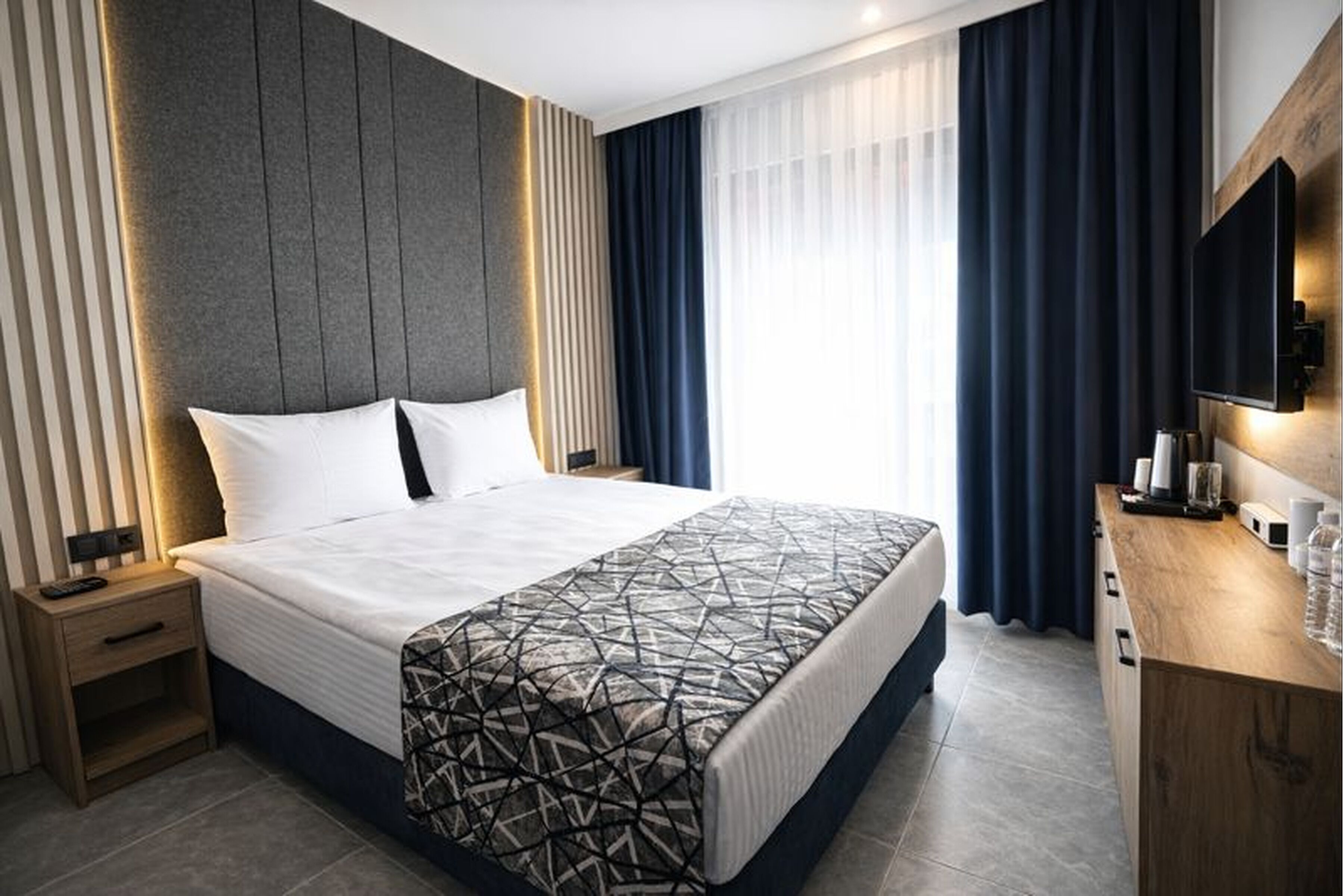 Ein modernes Hotelzimmer mit einem großen Bett, einem Nachttisch mit Lampe, einer Holzkommode und einem an der Wand befestigten Flachbildfernseher.