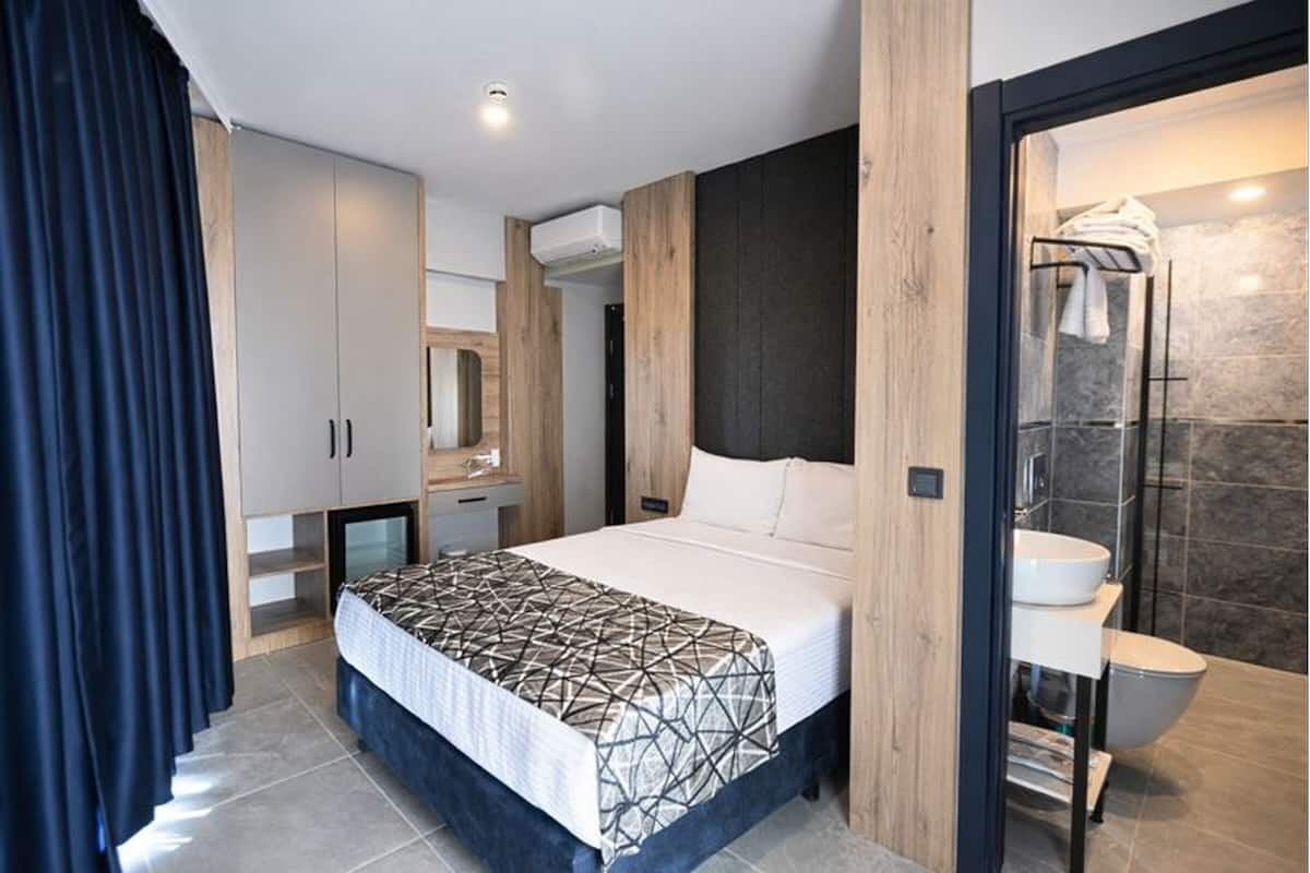 Una habitación de hotel moderna con una cama grande, un armario empotrado, un baño con cabina de ducha de vidrio y una pequeña zona de escritorio.