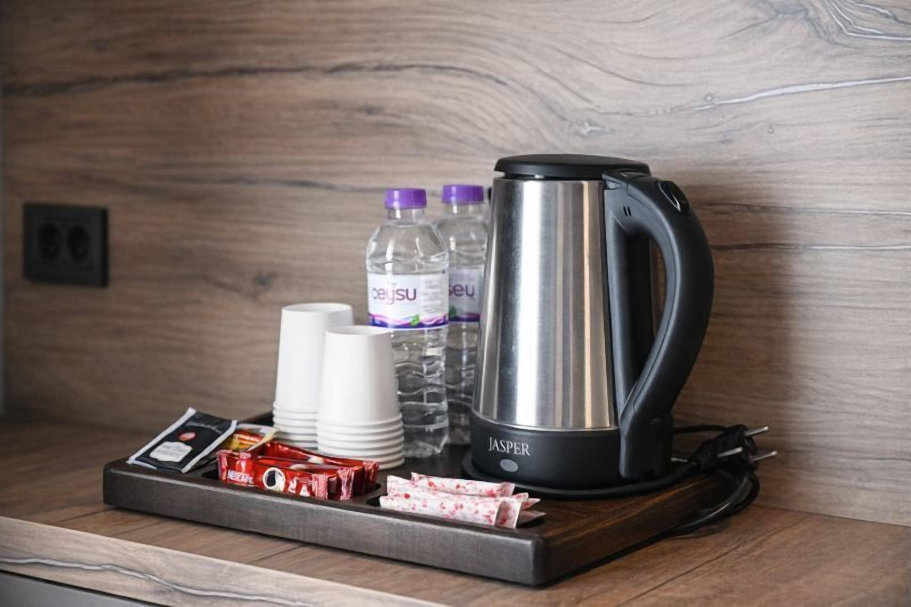 A coffee station with a black electric kettle, white cups, water bottles, and a tray with coffee pods and a small box.