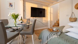 Ett modernt rum med ett glasbord, stolar, en säng med kuddar, en tv och en vas med blommor.