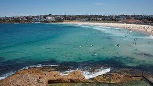 A coastal beach with clear waters, a rocky shoreline, and a cityscape in the background. - Bondi Waterfront Pad (North Bondi)
