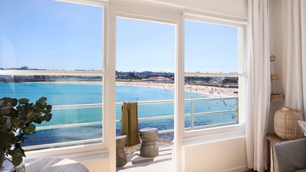 A room with large windows overlooking a beach, a small stool, and a wicker basket. - Bondi Waterfront Pad (North Bondi)