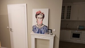Une pièce avec une porte blanche, une cheminée blanche surmontée d’un tableau encadré, et un coin cuisine avec des meubles blancs et une cuisinière à l’arrière-plan.