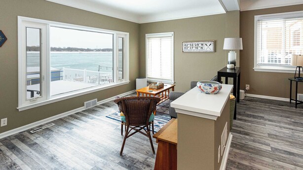 A room with a large window overlooking a body of water, a dining area with a table and chairs, and a small kitchen area with a counter and a lamp. - Serene Waterfront Getaway | Patio and Dock (Marysville)