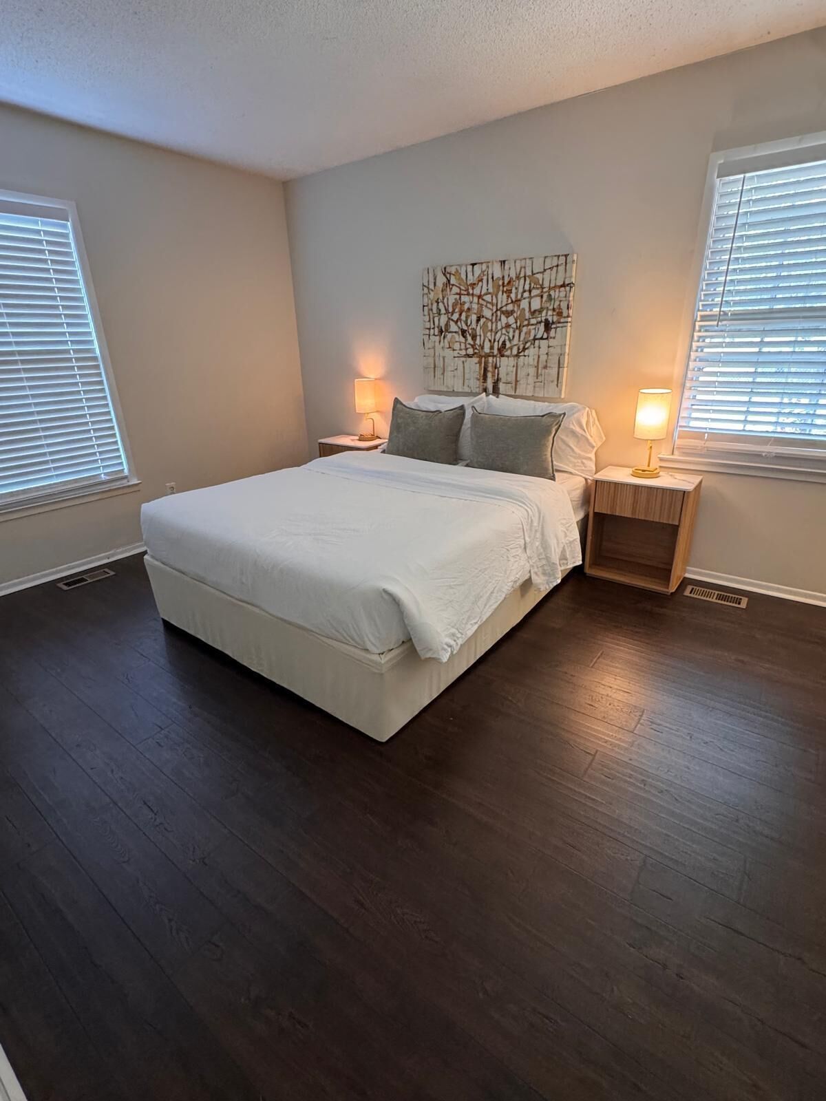A bedroom with a large bed, two bedside tables with lamps, a wall art piece, and a window with blinds.