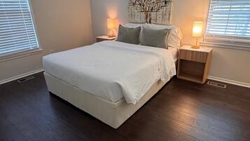 A bedroom with a large bed, two bedside tables with lamps, a wall art piece, and a window with blinds.
