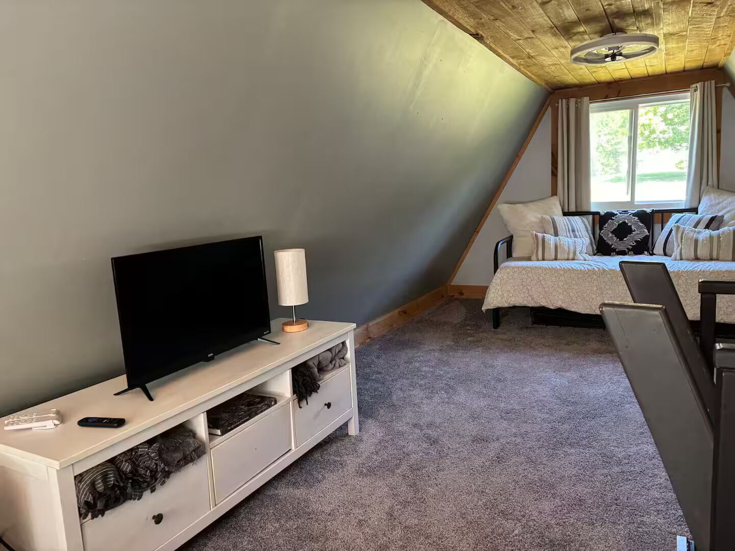 Un dormitorio con televisor sobre una consola blanca, cama con cubrecama blanco y beige, y ventana con cortinas.
