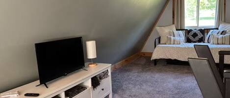 Un dormitorio con televisor sobre una consola blanca, cama con cubrecama blanco y beige, y ventana con cortinas.