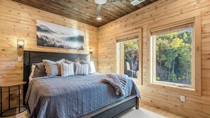 Ein Schlafzimmer in einer Holzhütte mit einem großen Bett, einem Waldgemälde und zwei Fenstern mit Blick ins Freie.