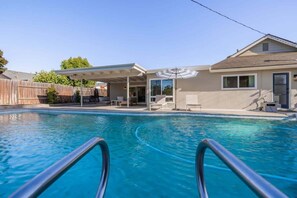 A swimming pool with a covered patio area, a wooden fence, and a house in the background. - @ Marbella Lane - Cozy Modern Home w/ Relaxing Design & Pool (Fullerton)