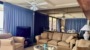 A living room with a beige sofa set, a glass coffee table, a flat-screen TV, and a ceiling fan. - Marbella Lane -  Beachfront  Haven w/ MT Views! (Waianae)