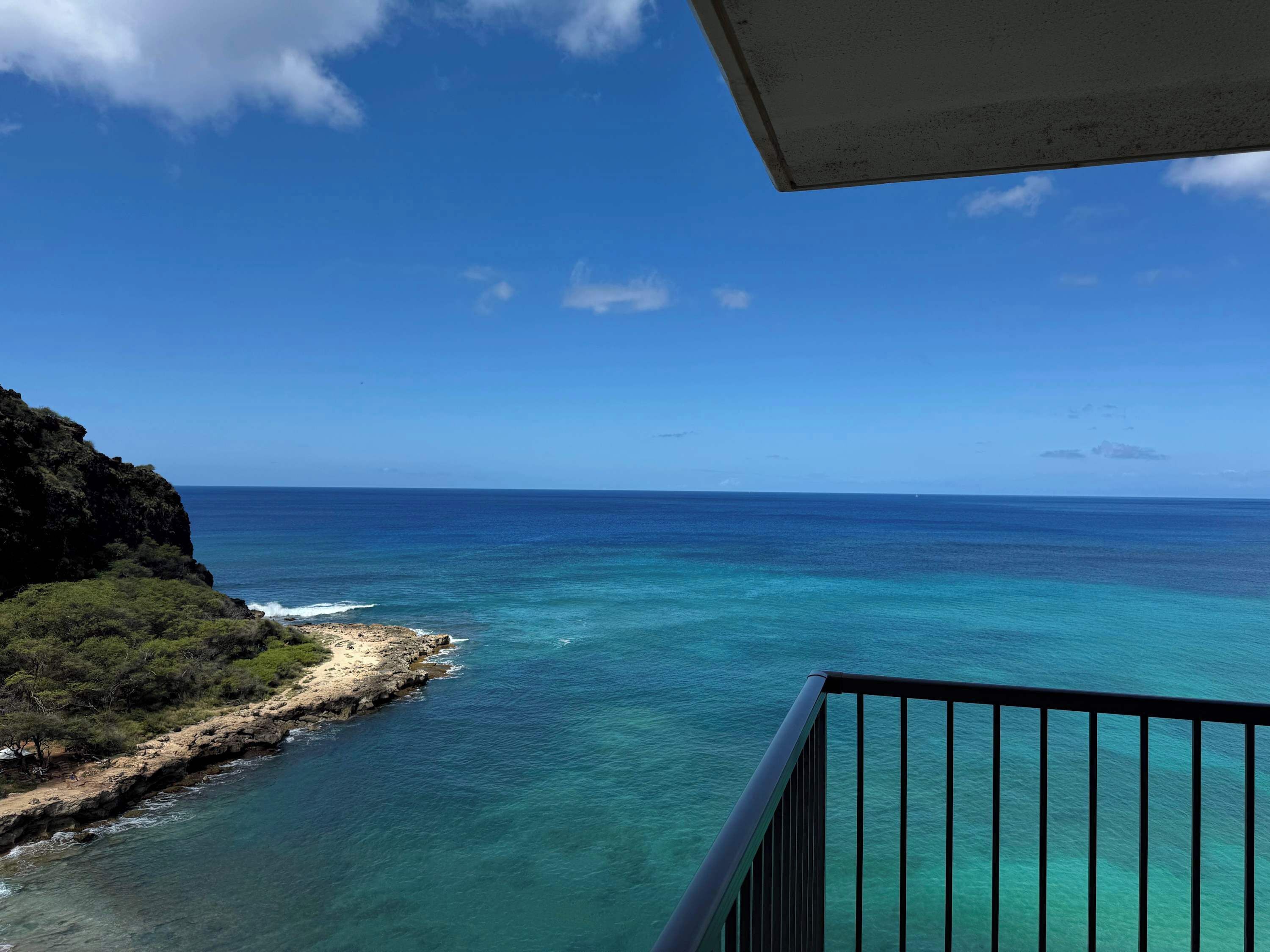 Udsigt over kysten fra en balkon med gelænder, hvor man kan se turkisblåt vand og en klippefyldt strand med sparsom vegetation.