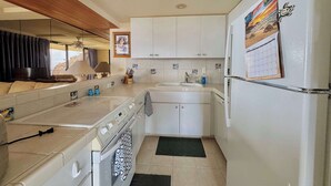 A compact kitchen with white cabinets, a built-in oven, a refrigerator, and a window overlooking a scenic view. - Marbella Lane -  Beachfront  Haven w/ MT Views! (Waianae)
