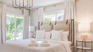 Una cama grande con sábanas blancas, una mesita de noche con lámpara y vistas al exterior con vegetación.