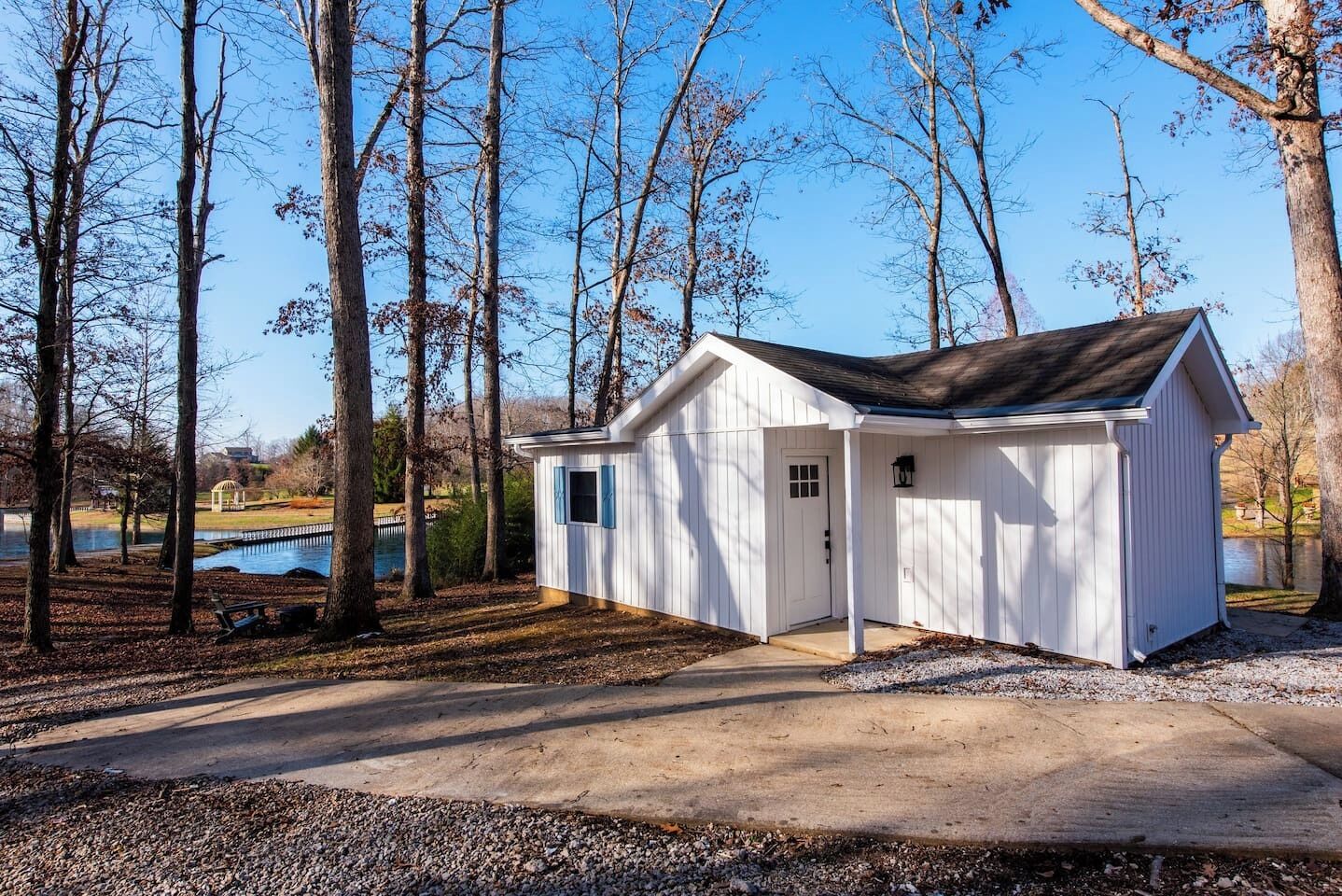 A small, single-story shed with a door and windows, surrounded by trees and a gravel path, near a body of water.
