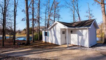 A small, single-story shed with a door and windows, surrounded by trees and a gravel path, near a body of water.