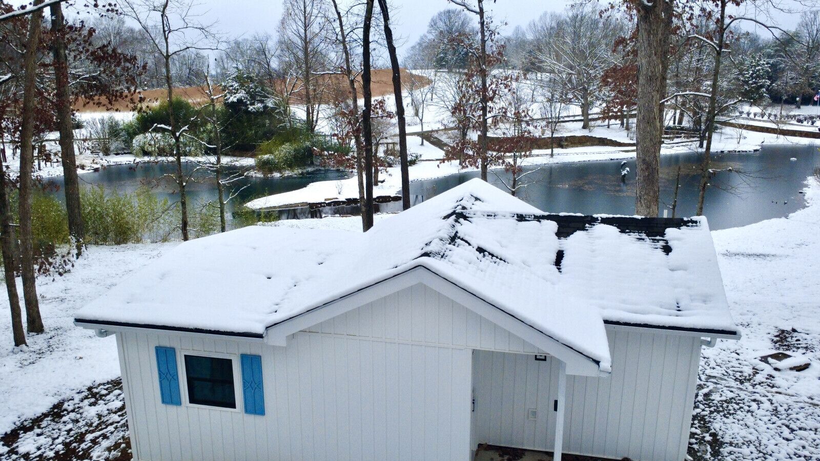 Un piccolo edificio bianco con tetto innevato e una finestra blu, circondato da alberi e un corso d'acqua.