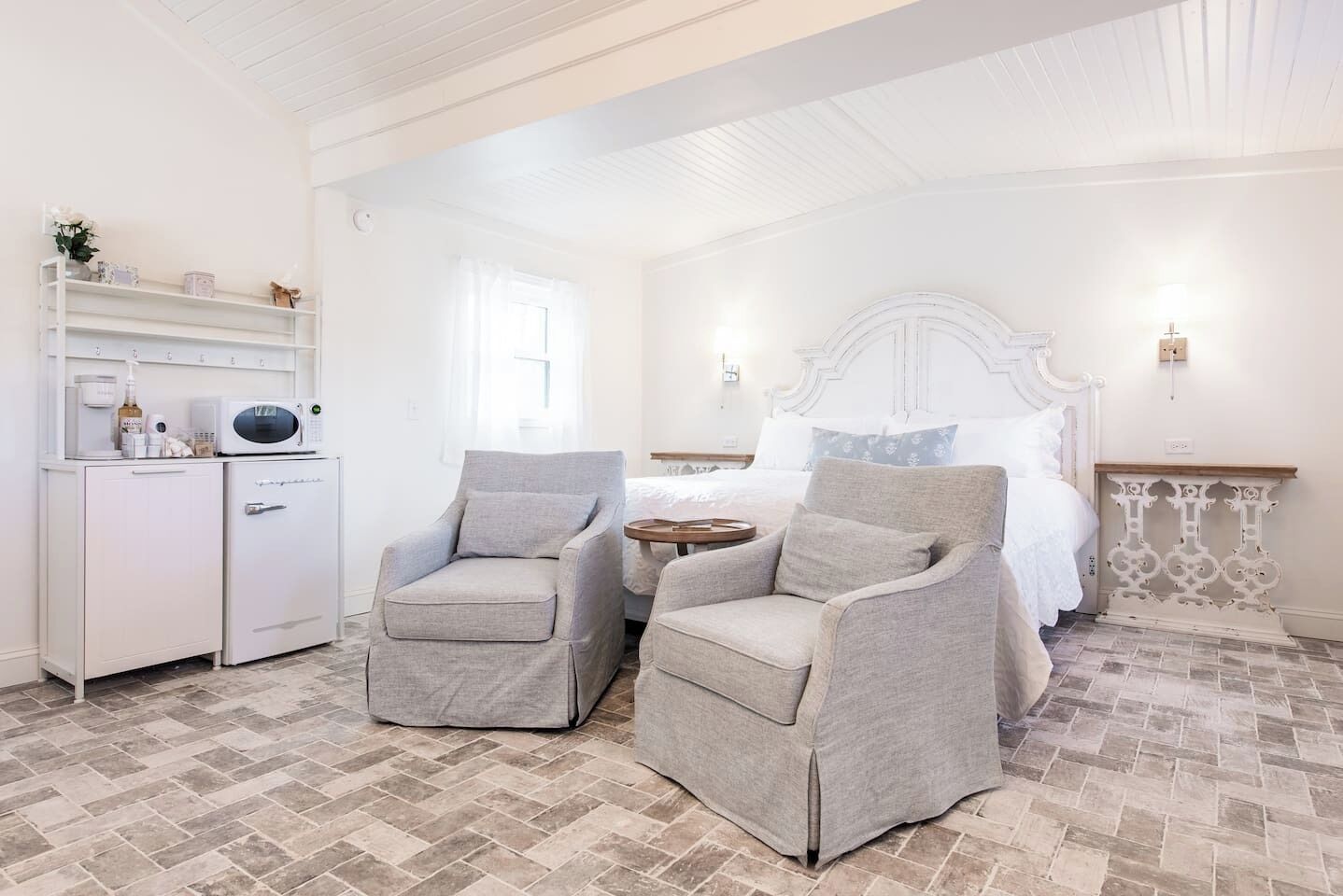 A bedroom with a bed, two armchairs, a small table, a white cabinet with a microwave, and a decorative wall piece.