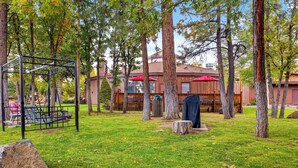 A grassy outdoor area with trees, a swing set, and a building with red awnings in the background. - Pine-Lodge by Roundhouse Resort and Spa (Pinetop Country Club)