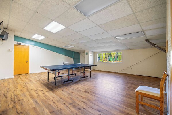 A room with a ping pong table, wooden flooring, a door, a window with a view, and a chair.