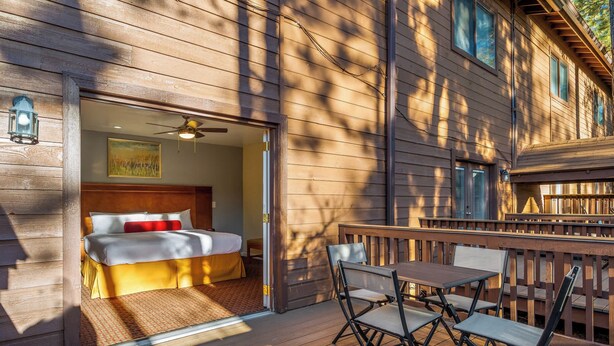 Uma cabana de madeira com um quarto, que inclui uma cama, um ventilador de teto e um quadro na parede. Lá fora, há um alpendre com uma mesa e cadeiras.
