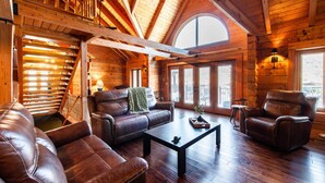 L’intérieur d’une cabane en rondins, avec un plafond haut, des poutres en bois, une cheminée, des meubles en cuir et de grandes fenêtres.