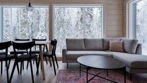 Ein modernes Interieur mit einem grauen Sofa, einem runden Couchtisch und schwarzen Stühlen. Große Fenster bieten Blick auf schneebedeckte Bäume.