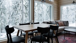 Ein gemütliches Interieur mit Esstisch und Stühlen, einem grauen Sofa und Blick auf verschneite Bäume durch große Fenster.