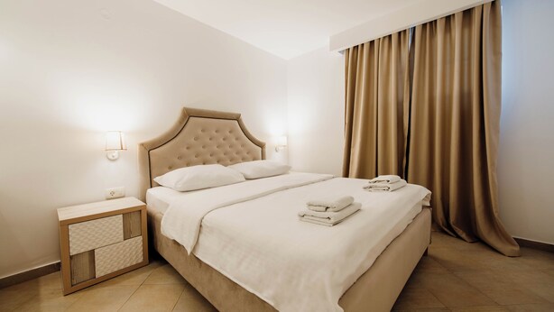 A neatly made bed with a headboard, a bedside table with a lamp, and a window with curtains. - Yellow 1-bedroom apartment with AC in vibrant Budva (Budva)