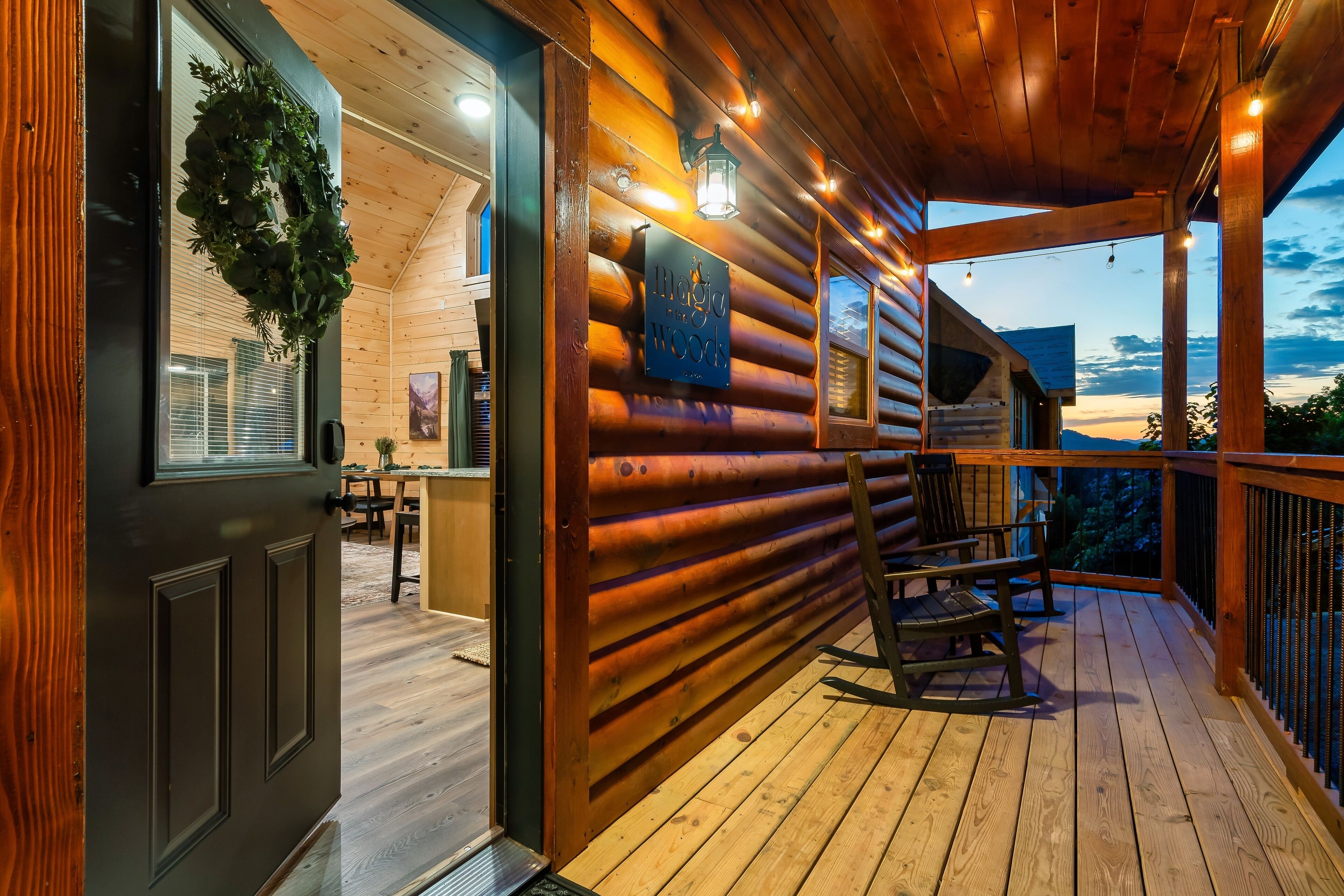 A log cabin with a wooden deck, a sign reading "Magic Woods," and a view of the mountains at dusk.