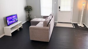 Een woonkamer met een beige bank, een flatscreen-tv op een witte standaard, twee ramen met jaloezieën, een potplant en een vloerlamp.