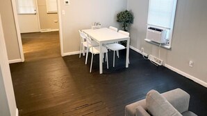 Een kamer met een witte tafel en stoelen, een grijze bank en een raam met jaloezieën.