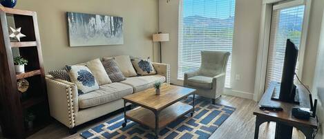 A living room with a sofa, coffee table, and armchair, a ceiling fan, a painting, and a window with blinds.
