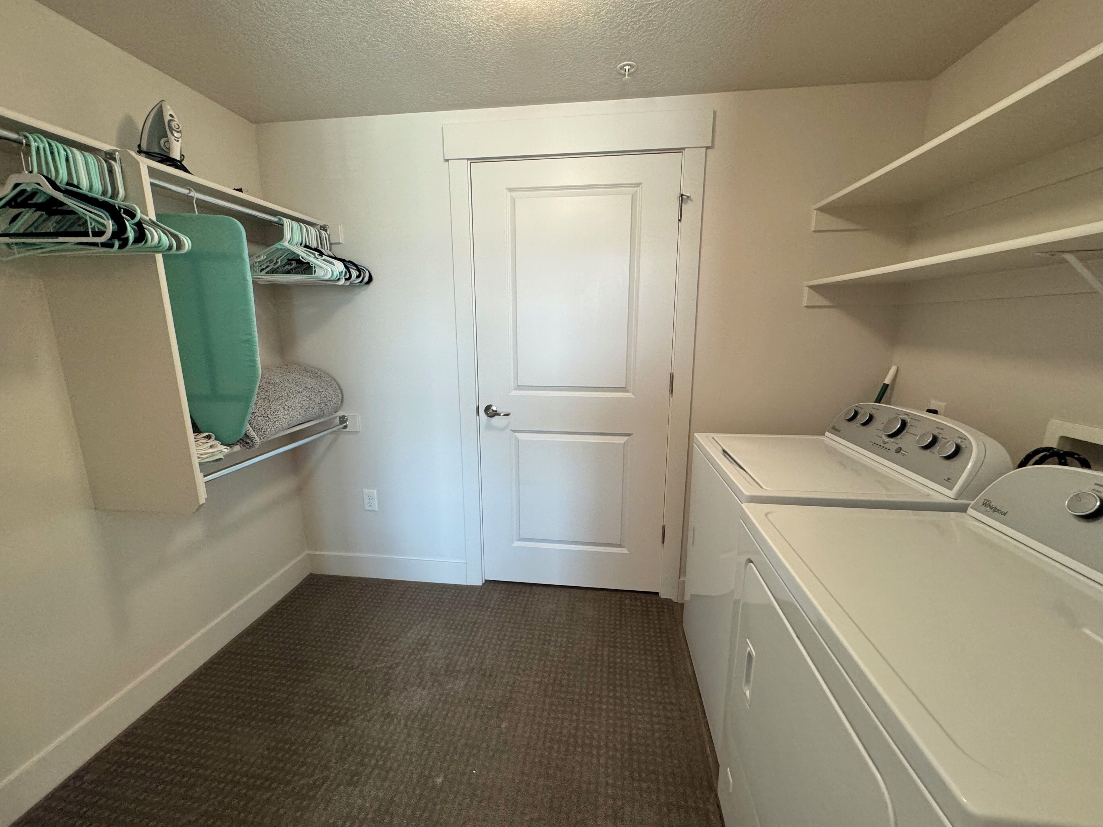A laundry room with a washer and dryer, a door, and shelves with hanging clothes.