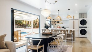 Una cuina moderna amb armaris blancs, una illa central, il·luminació penjant i vistes a una zona de piscina exterior.