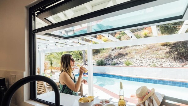 Habitació amb finestra gran que dona a una piscina, taula de menjador amb menjar i begudes, i una cadira.