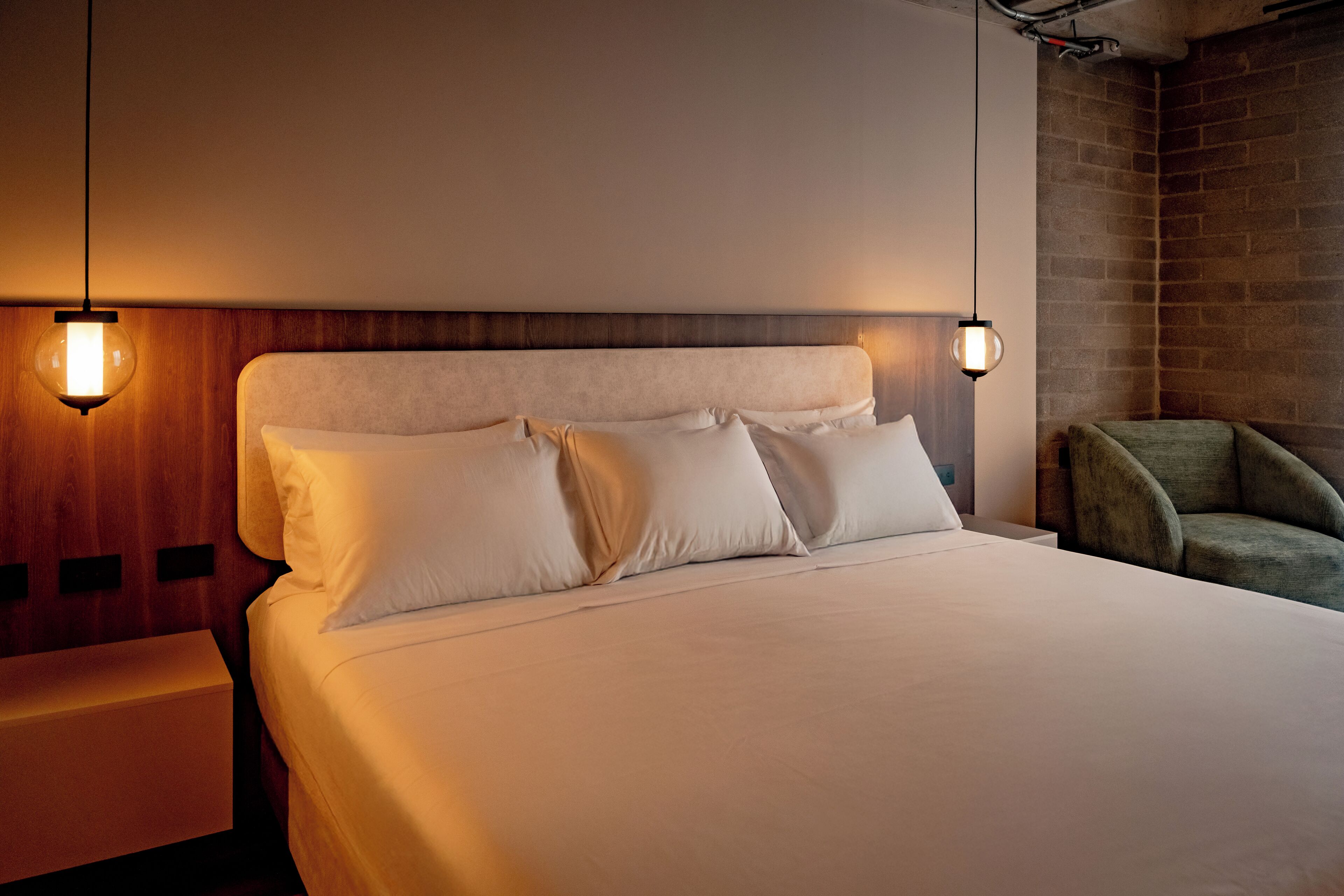 Una cama bien hecha con sábanas y almohadas blancas, un cabecero de madera y dos lámparas colgantes.