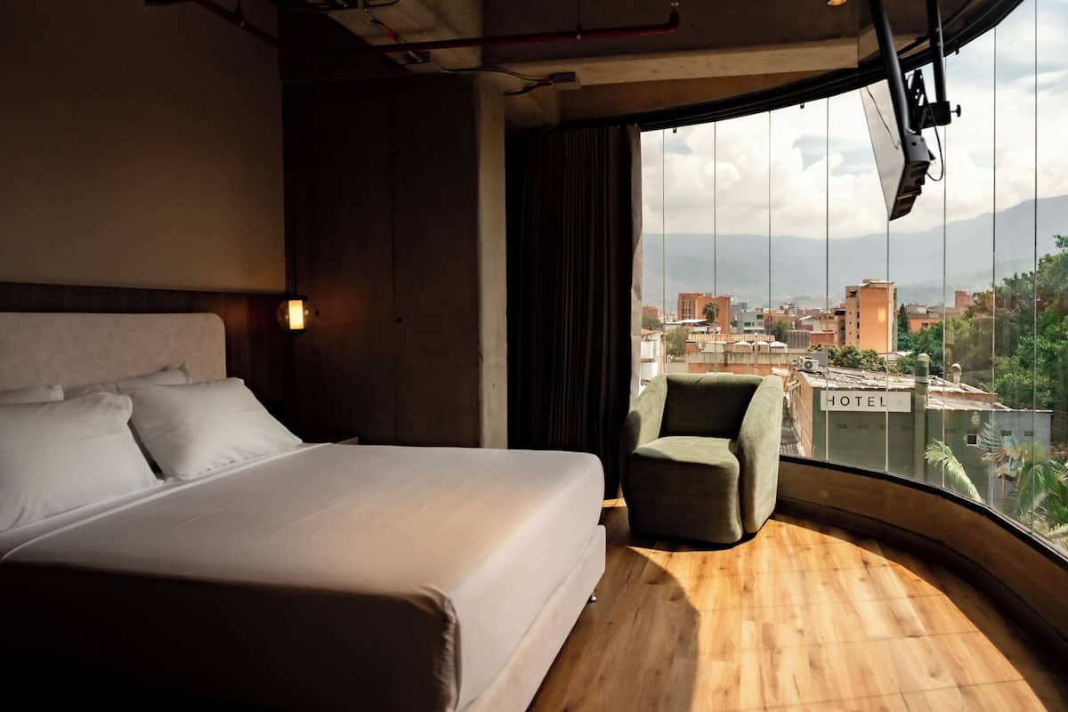 Ein Hotelzimmer mit einem großen Bett, einem kleinen Sessel und Blick auf die Stadt durch ein großes Fenster.