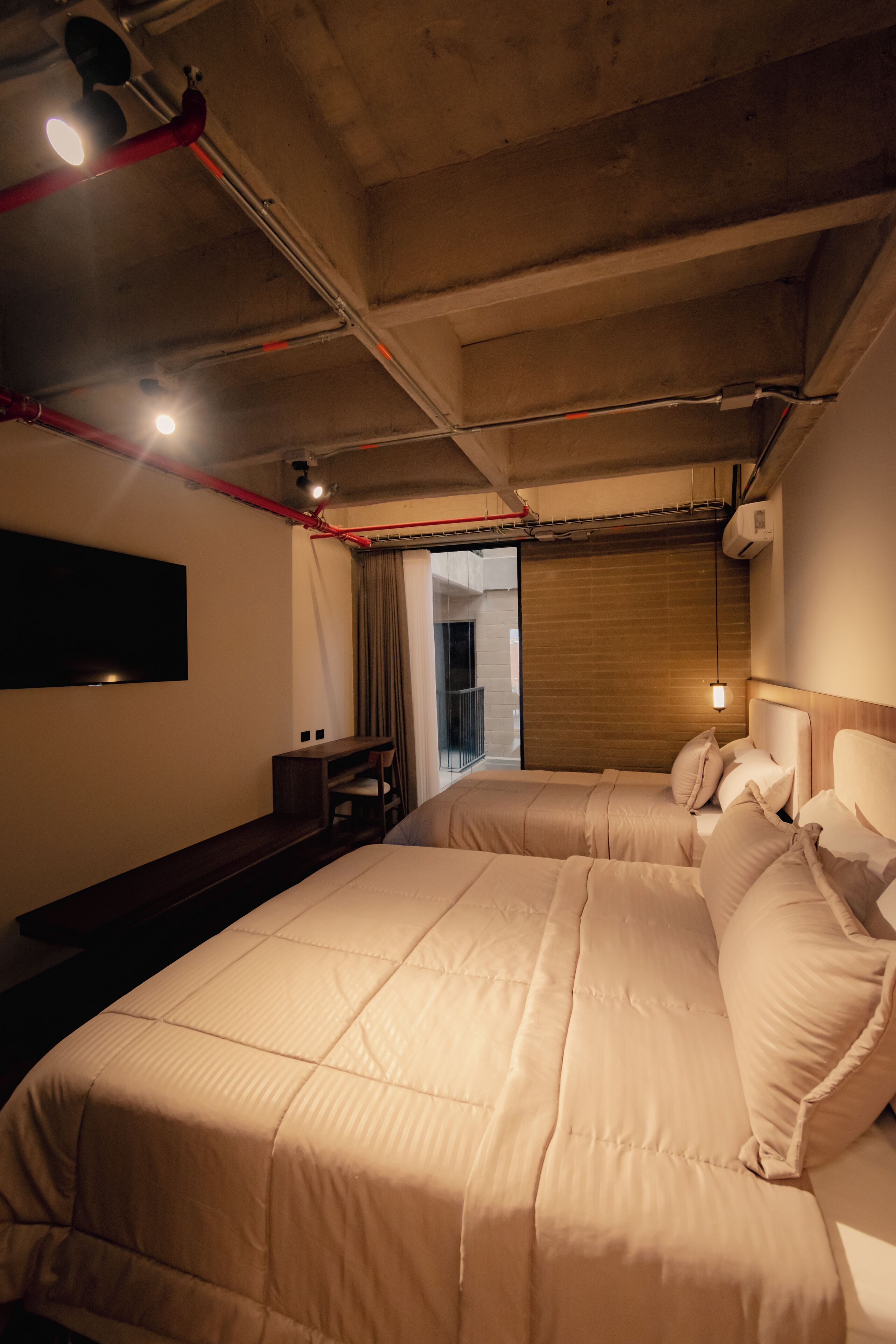 A hotel room with two beds, a flat-screen TV mounted on the wall, a wooden headboard, and a window with curtains.