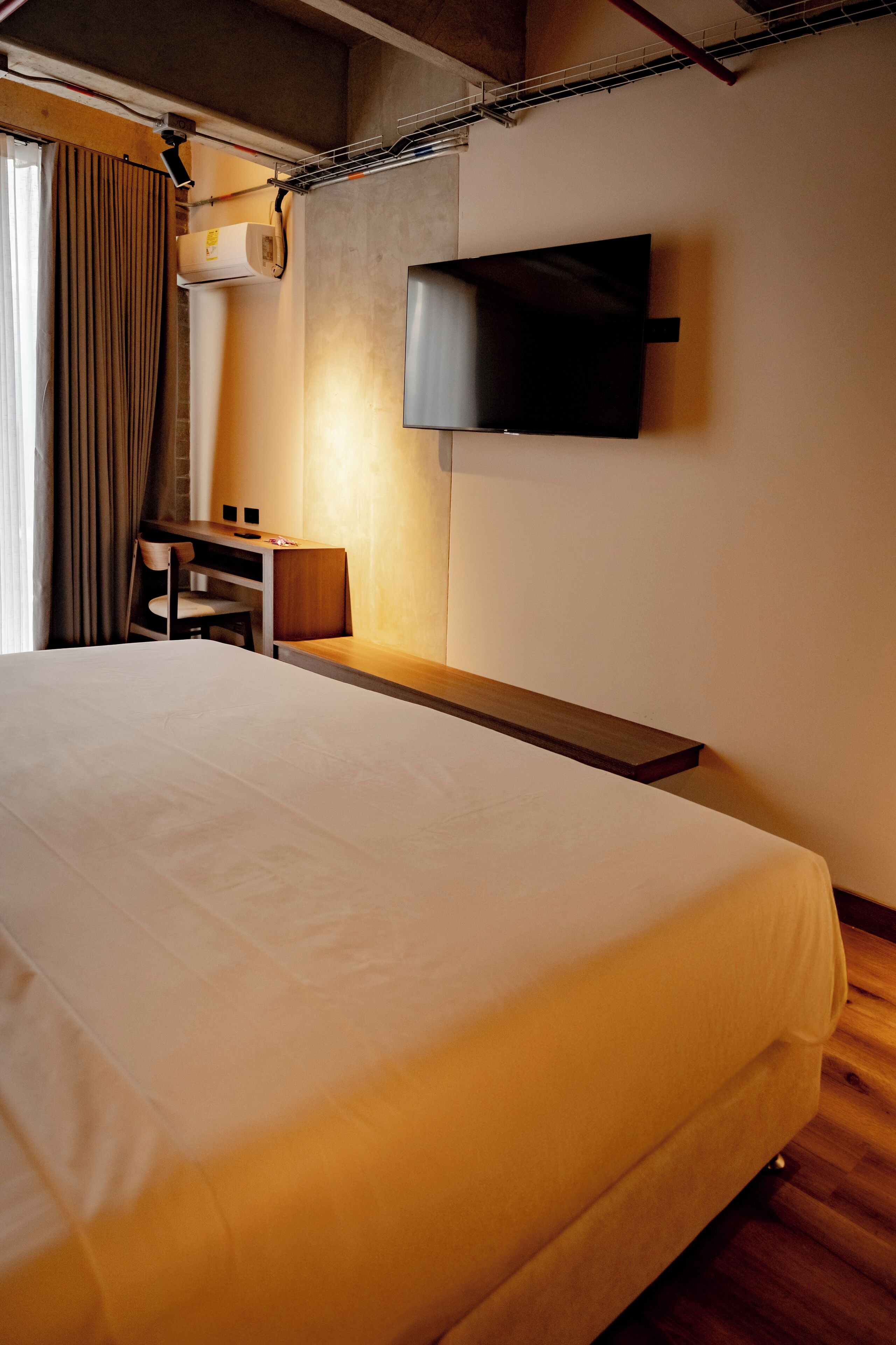 A hotel room with a large bed, a desk, a chair, a flat-screen TV mounted on the wall, and an air conditioning unit.