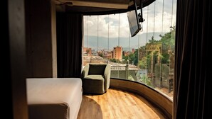 Habitación de hotel con piso de madera curvada, un sillón verde individual y un amplio ventanal con vista a la ciudad.