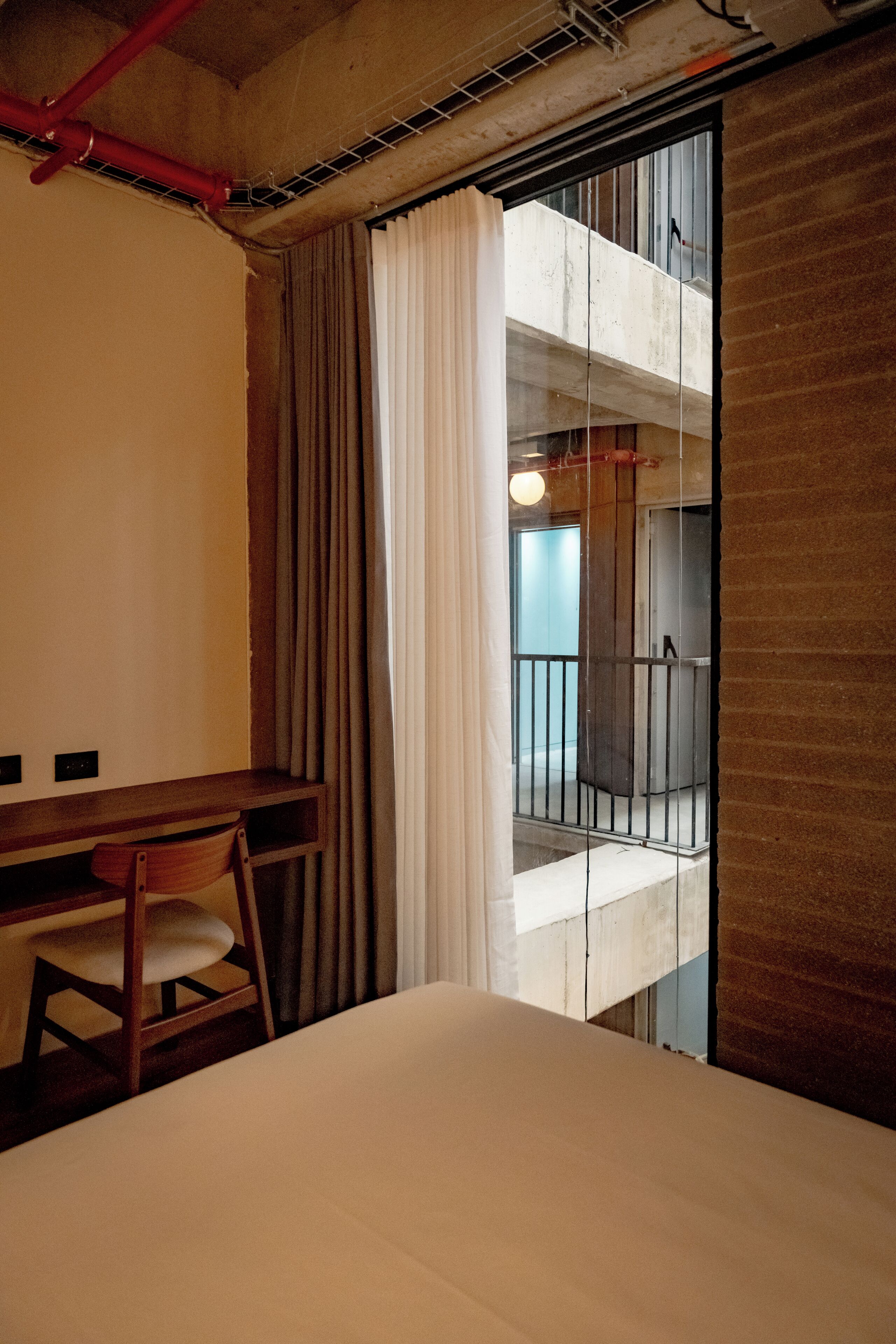 A hotel room with a bed, a wooden desk, a chair, and a view of a balcony with a railing.