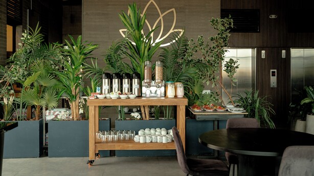 Un interior moderno con una mesa de madera que exhibe diversos objetos, rodeado de plantas verdes y una pared con diseño geométrico.
